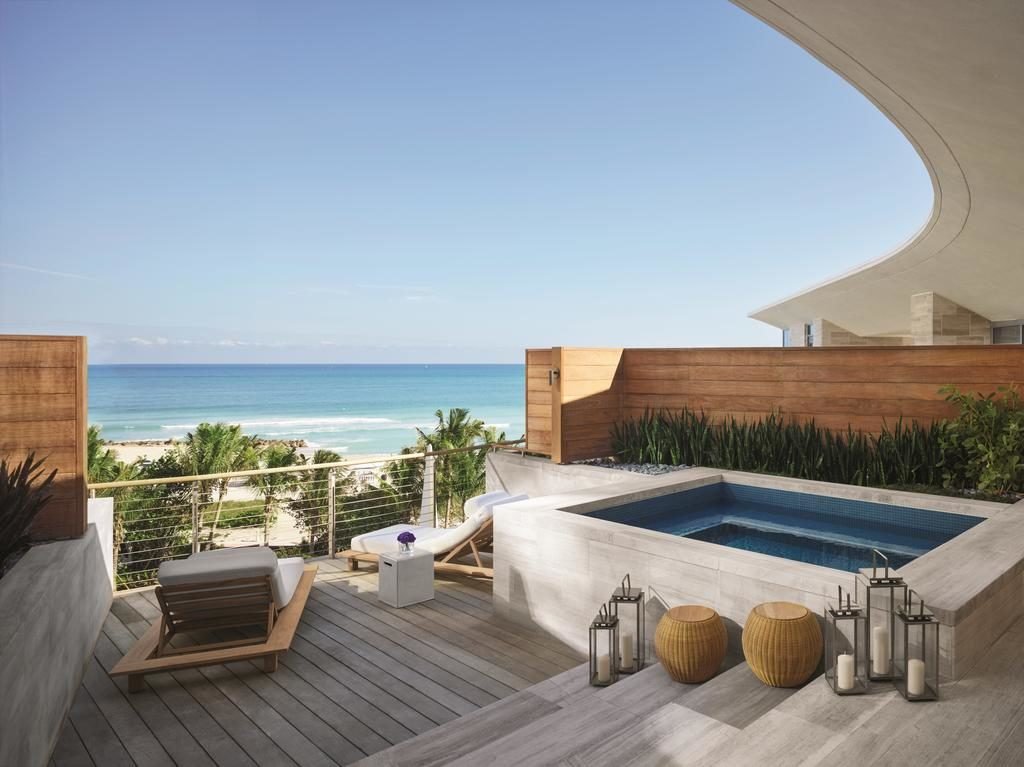 A imagem mostra um terraço luxuoso com vista para o mar no The Miami Beach EDITION. O espaço inclui uma pequena piscina de hidromassagem embutida no chão, rodeada por um deck de madeira. Há uma espreguiçadeira e uma mesa lateral, perfeitas para relaxar. Lanternas decorativas e cestos estão dispostos ao redor da piscina. A cerca de madeira proporciona privacidade, enquanto plantas verdes acrescentam um toque natural. Ao fundo, vê-se a praia e o oceano, com palmeiras complementando a vista serena e tropical.