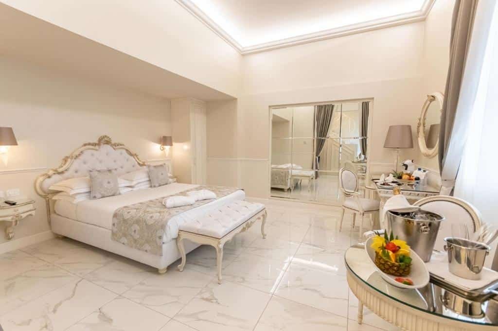 Quarto deluxe do The Moon Boutique Hotel & Spa, de 40 m², com decoração intimista e elegante utilizando cores claras (branco e bege), além de cama de casal, mesa com cadeira, espelho grande, e mesa redonda com frutas, balde de gelo e balde de taça em cima.