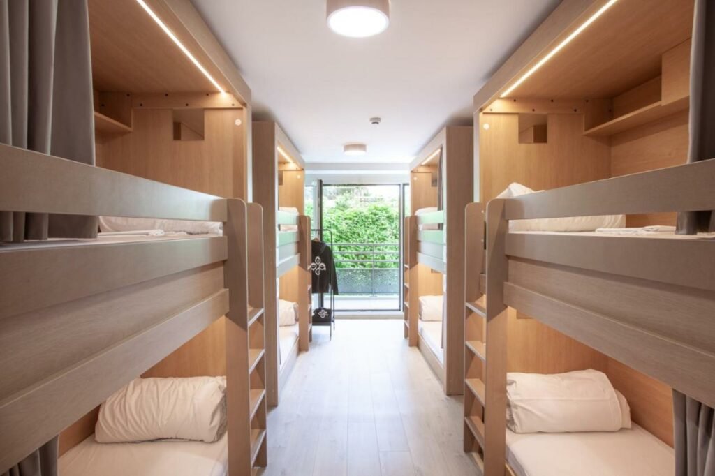 Quarto do The People - Paris Bercy. Beliches bege ladeiam a imagem enquanto o corredor no centro leva até uma janela de vidro com vista para árvores. Há um cabide de roupas ao lado esquerdo da janela.