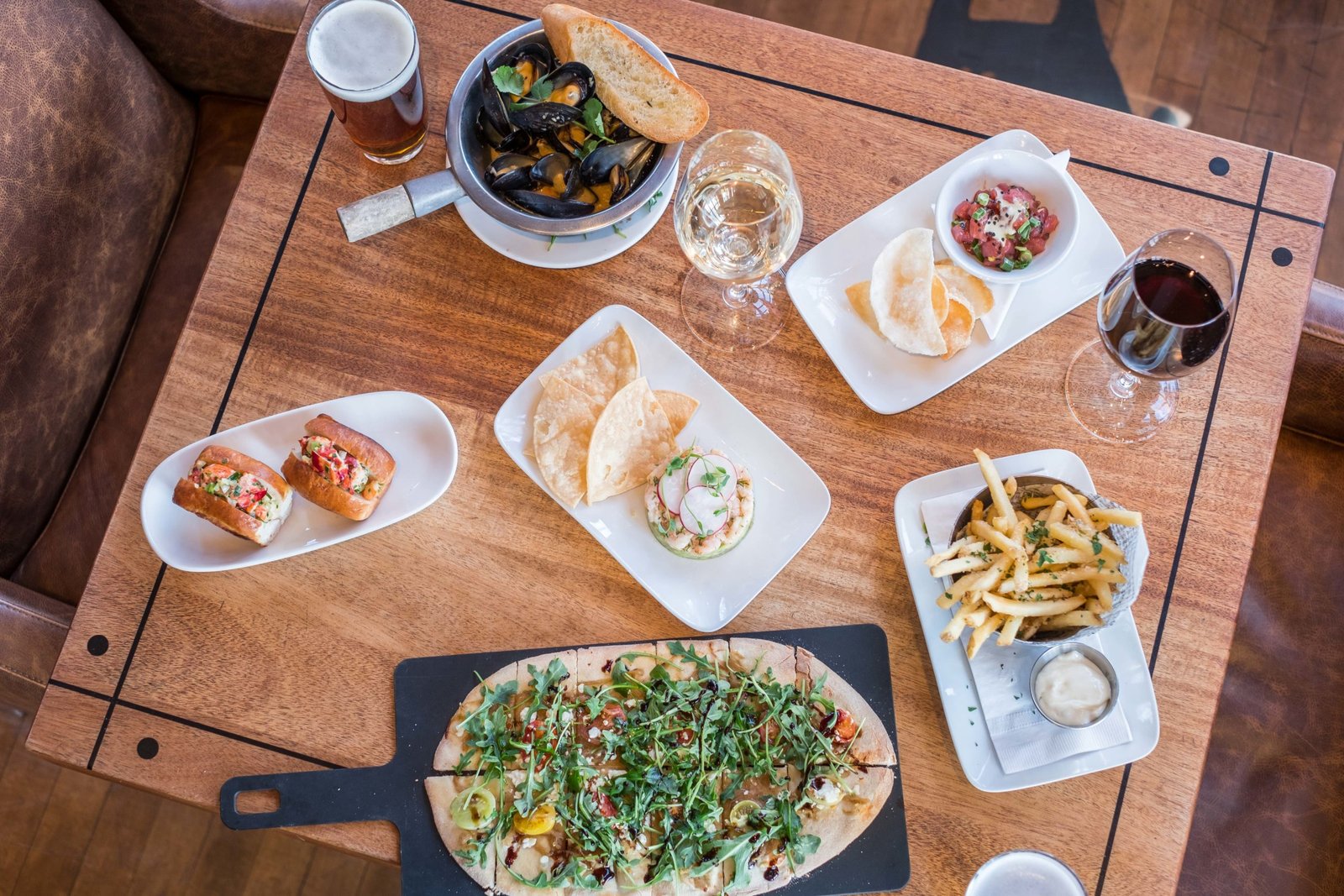 Uma mesa de madeira com uma variedade de pratos, incluindo pão achatado, batatas fritas, tacos, mexilhões, batatas fritas com molho e taças de vinho e cerveja no The Sandbar Seafood Restaurant.