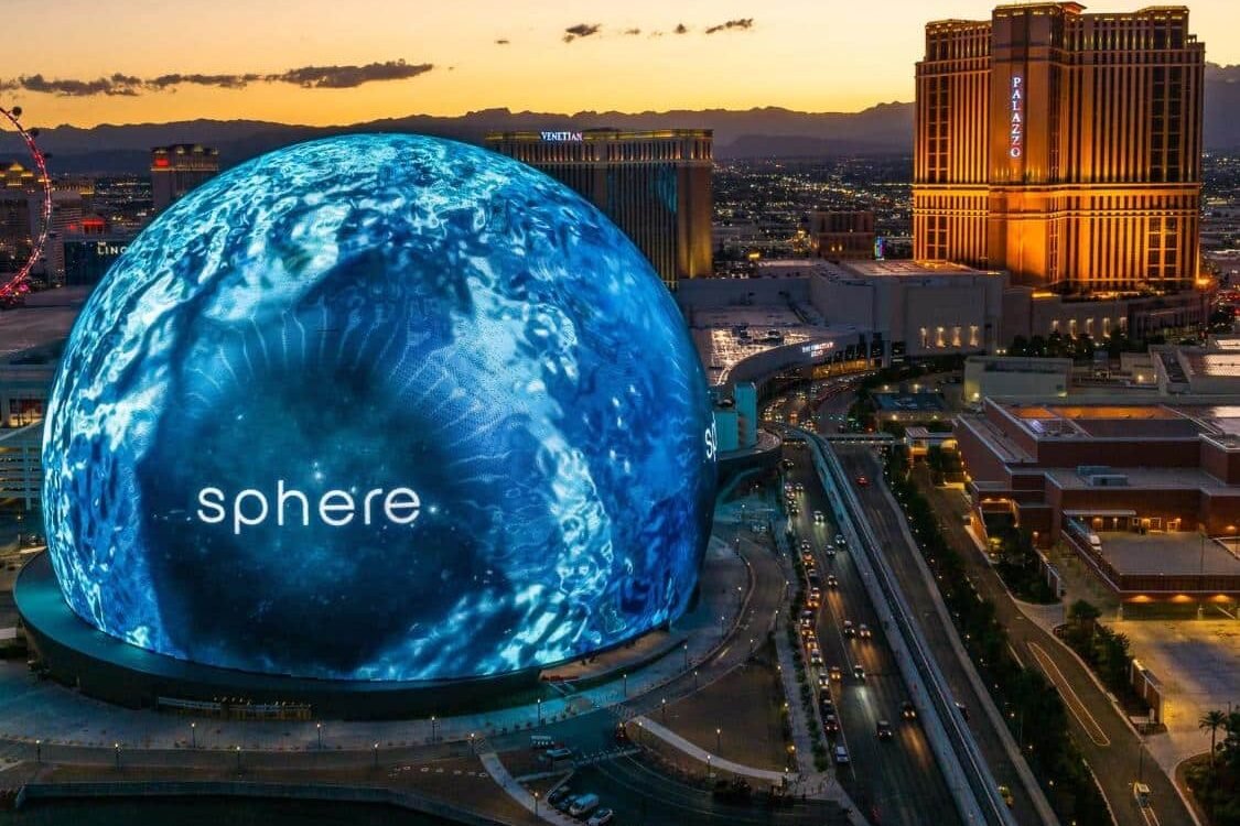 A The Sphere, esfera gigante, estão ao lado esquerdo da foto. Ela está toda azul, apenas com a palavra "sphere" escrita. Do lado direito estão a avenidade e alguns hotéis.