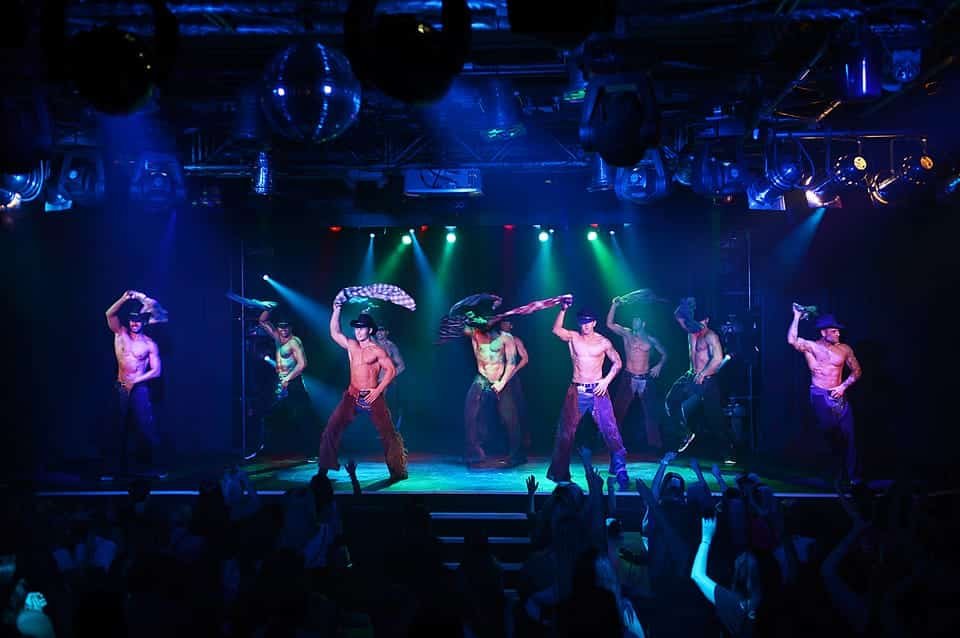 Seis artistas masculinos do Thunder From Down Under. Eles estão no palco, sem camisa, usando chapéus e calças de cowboy, segurando camisas acima da cabeça. Público em primeiro plano.