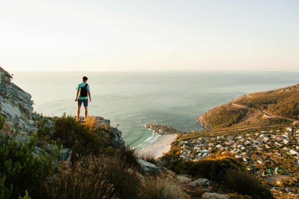 Homem de costas na beira do precipício enquanto olha para casas à beira-mar na Cidade do Cabo. O local é repleto de vegetação nativa e tem pedras na trilha. - Foto: Tim Johnson via Unsplash