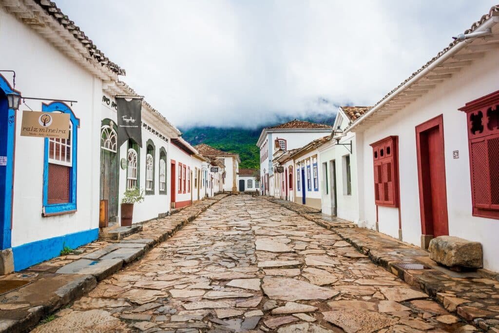 Uma rua estreita de paralelepípedos é ladeada por pequenos edifícios coloridos em estilo colonial sob um céu nublado.