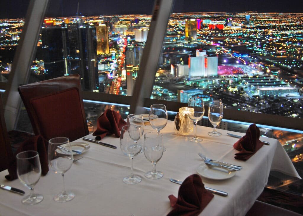 Vista de uma das mesas do restaurante Top of the World durante à noite. A parede é toda de vidro, então é possível ver todos os edifícios e avenidas iluminadas lá embaixo.