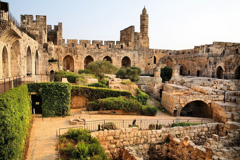 Antiga fortaleza da Torre de David em Israel feita de pedra com paredes arqueadas, uma torre e vegetação exuberante. Ruínas e caminhos são visíveis no primeiro plano.