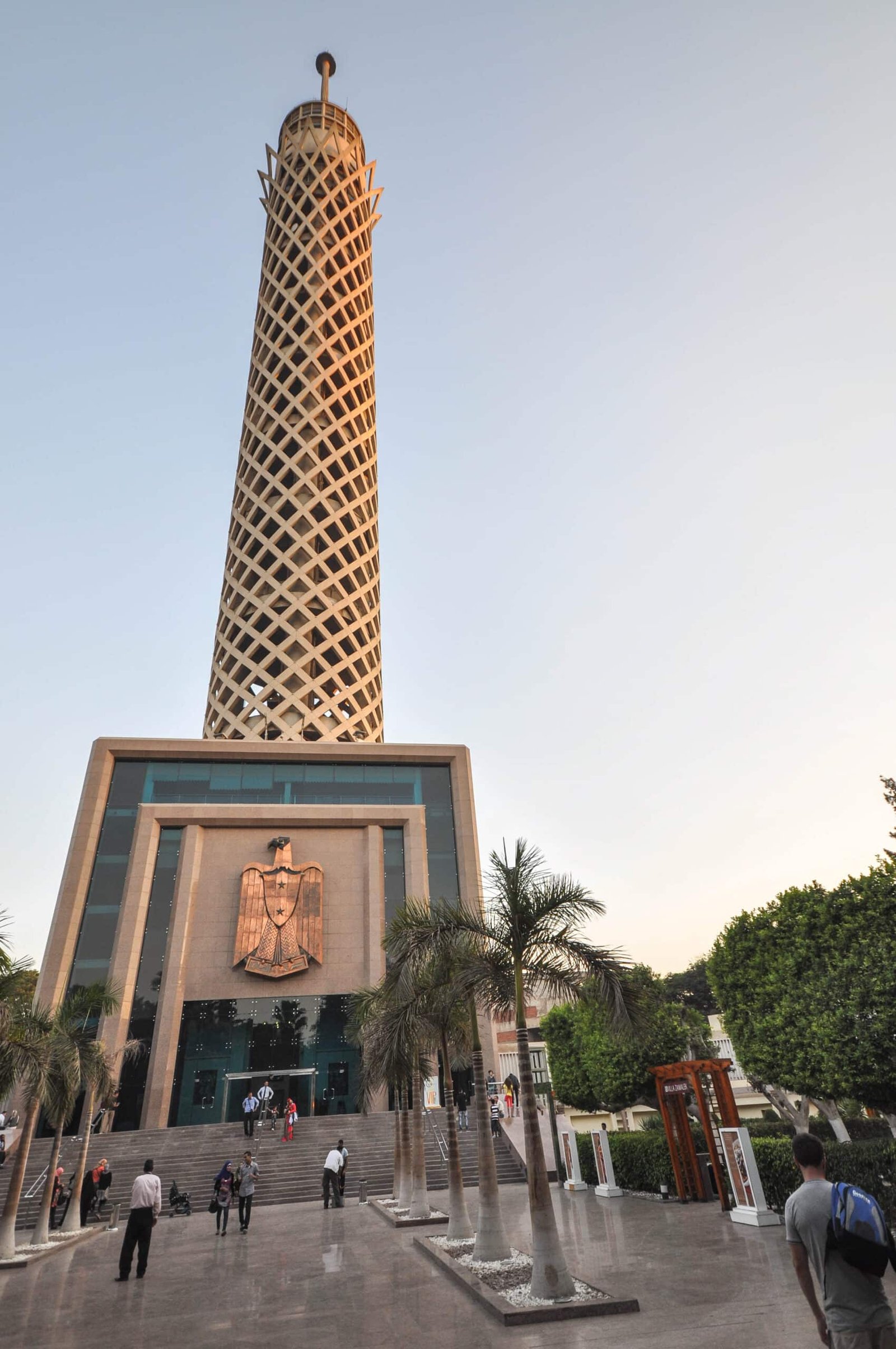 A Torredo Cairo em estilo treliça, com uma base larga, apresentando um emblema. As pessoas andam e se reúnem ao redor da entrada, ladeada por árvores.