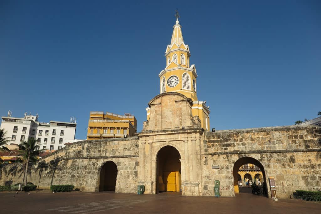 A imagem mostra uma entrada imponente de uma cidade murada, Cartagena, destacando um grande arco de pedra no centro, flanqueado por dois arcos menores. Acima do arco central, há uma torre amarela com detalhes brancos e um relógio grande. A torre é alta e esbelta, com uma arquitetura que se destaca contra o céu azul claro. À esquerda da imagem, há edifícios modernos de cores claras.