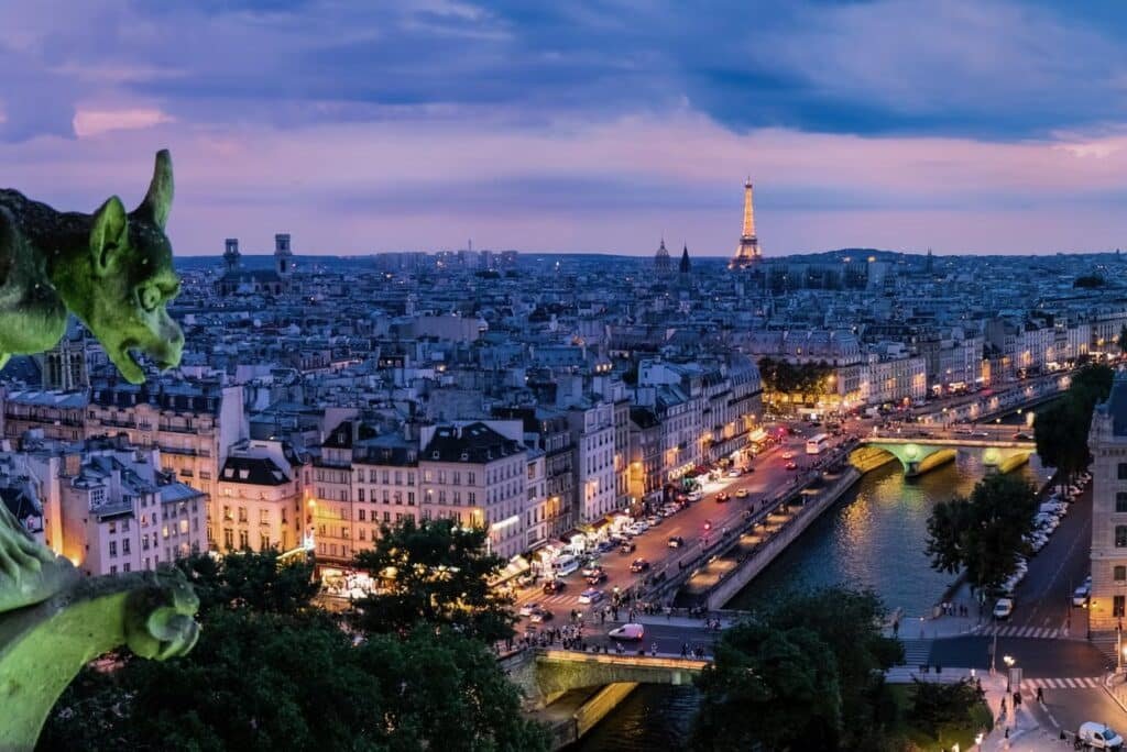 Gárgula com vista para uma paisagem urbana ao anoitecer, com um rio, ruas iluminadas e uma torre proeminente ao longe. A foto ilustra o post sobre passeios em Paris.
