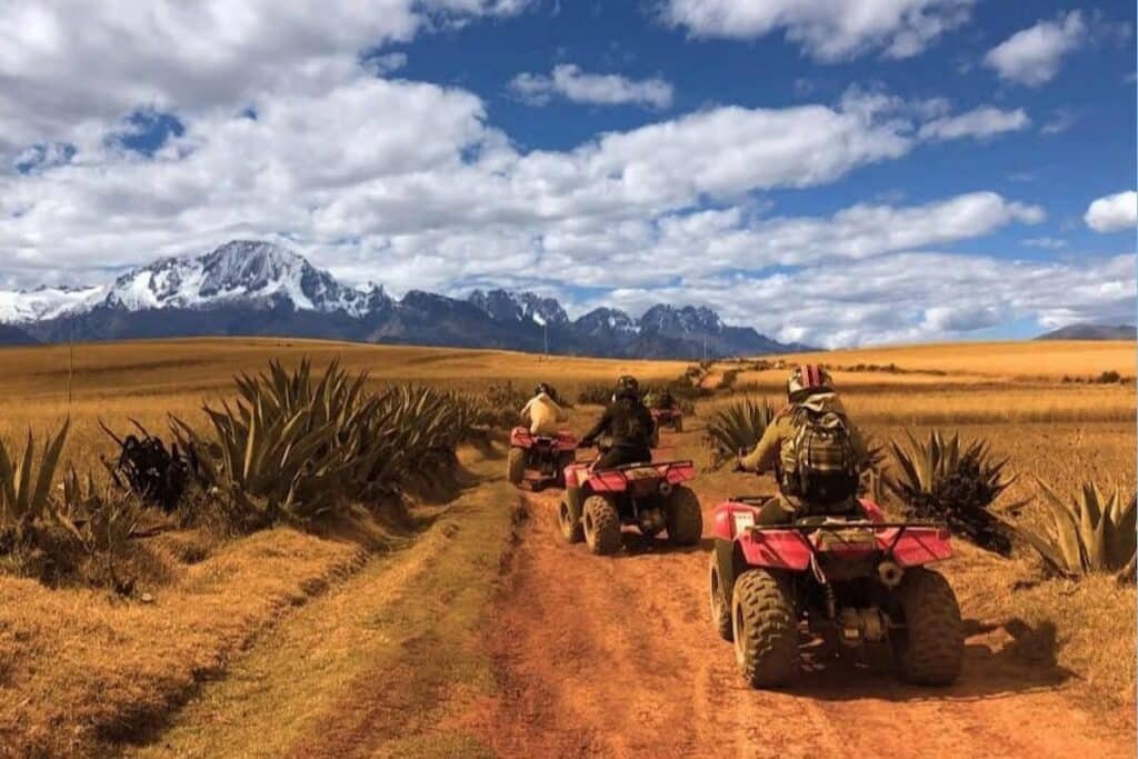 três carrinhos de quadriciclo com mulheres sentadas em cima dirigindo, de costas para a câmera. Elas estão numa estrada de terra com vista para as montanhas peruanas, onde os topos estão cheios de neve, e o céu está azul e com poucas nuvens brancas e fofas.