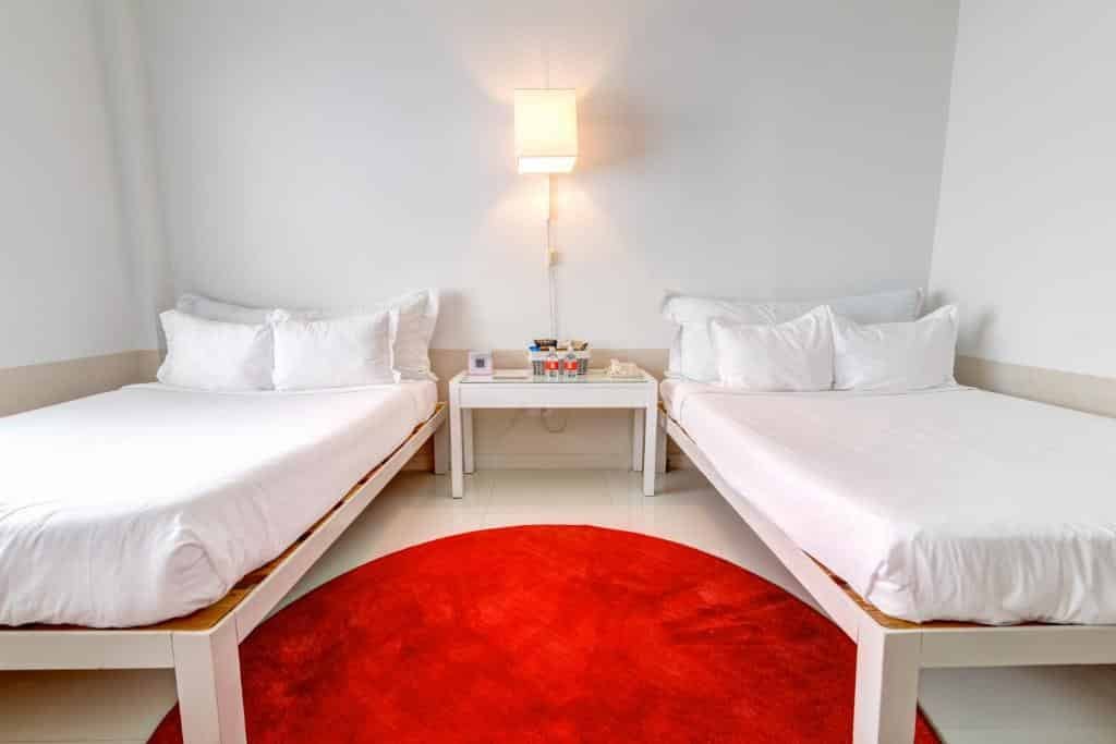 A imagem mostra um quarto simples e moderno com duas camas de solteiro no Townhouse Miami Beach. As camas têm roupa de cama branca, criando um ambiente limpo e minimalista. Entre as camas, há uma mesa de cabeceira branca com alguns itens, como um telefone e livros. Acima da mesa, uma lâmpada de parede ilumina o espaço. No chão, há um tapete redondo vermelho que adiciona um toque de cor vibrante ao ambiente predominantemente branco. As paredes são brancas, reforçando a sensação de simplicidade e clareza no design do quarto.