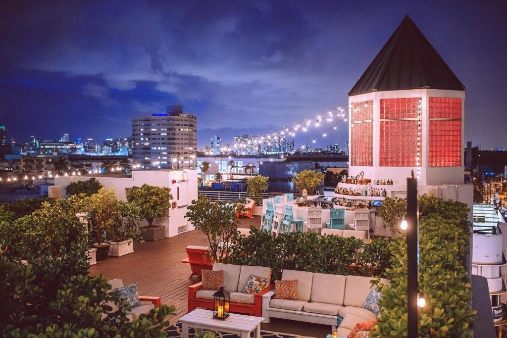 A imagem mostra um terraço iluminado à noite com uma vista panorâmica da cidade. O terraço tem uma área de estar com sofás e mesas decoradas com almofadas coloridas e lanternas. Há várias plantas ao redor, criando um ambiente aconchegante. Um bar ao fundo tem bancos altos em tons de branco e azul, e está decorado com luzes festivas. À direita, há uma torre com janelas vermelhas iluminadas. Luzes penduradas atravessam o terraço, adicionando um charme extra à cena noturna. Ao fundo, a silhueta dos prédios da cidade completa a vista deslumbrante. O terraço fica no Townhouse Miami Beach, um dos hotéis em Miami Beach indicados.