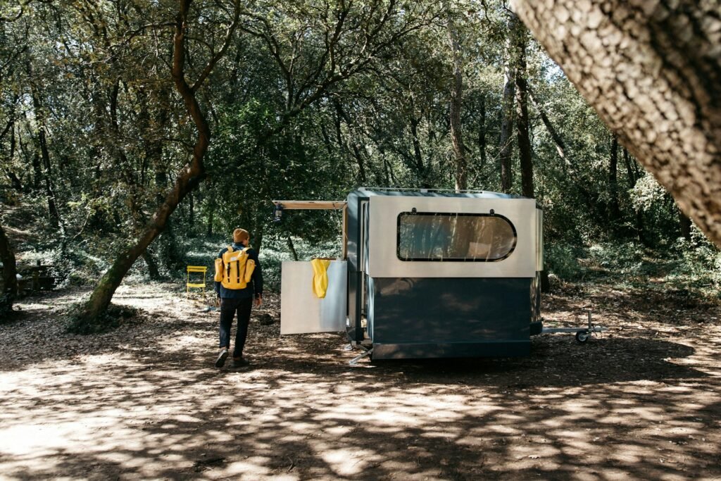 Homem de costas com uma mochila amarela nas costas, ao lado de um trailer que é rodeado de natureza
