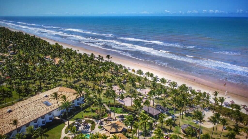 A imagem mostra uma vista aérea do Transamerica Comandatuba All Inclusive Resort, um dos melhores resorts all inclusive no Brasil. A paisagem exibe a extensa praia de areia clara ao lado direito da imagem, com ondas suaves do mar azul. À esquerda, o resort é cercado por uma vasta área verde com numerosas palmeiras, proporcionando um cenário tropical exuberante. Os edifícios do resort têm telhados de telha e estão intercalados com áreas de lazer e estruturas menores. A praia é equipada com guarda-sóis e espreguiçadeiras, e algumas pessoas caminham ao longo da orla, aproveitando o ambiente tranquilo e paradisíaco.