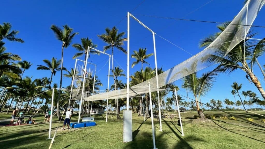 A imagem mostra uma estrutura de trapézio ao ar livre em meio a um cenário tropical, repleto de palmeiras e céu azul no Transamerica Comandatuba - All Inclusive Resort. A estrutura de metal tem redes de segurança abaixo e várias cordas e plataformas, sugerindo que é usada para atividades acrobáticas. Algumas pessoas estão presentes ao redor, observando ou se preparando para participar da atividade. O ambiente verdejante e a presença de palmeiras ao fundo conferem um aspecto relaxante e tropical à cena.