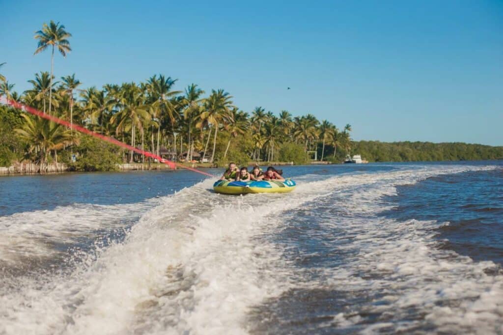 A imagem mostra um grupo de pessoas se divertindo em um bote inflável puxado por um barco em alta velocidade em um ambiente aquático. O bote, colorido em verde e azul, está sendo puxado por uma corda vermelha e desliza sobre as ondas criadas pelo barco. O cenário ao redor é repleto de palmeiras altas, criando um ambiente tropical e paradisíaco. O céu está limpo e azul, refletindo um dia ensolarado e ideal para atividades aquáticas. Ao fundo, pode-se ver a vegetação densa e uma linha costeira tranquila, sugerindo um local ideal para recreação e lazer no Transamerica Comandatuba - All Inclusive Resort.