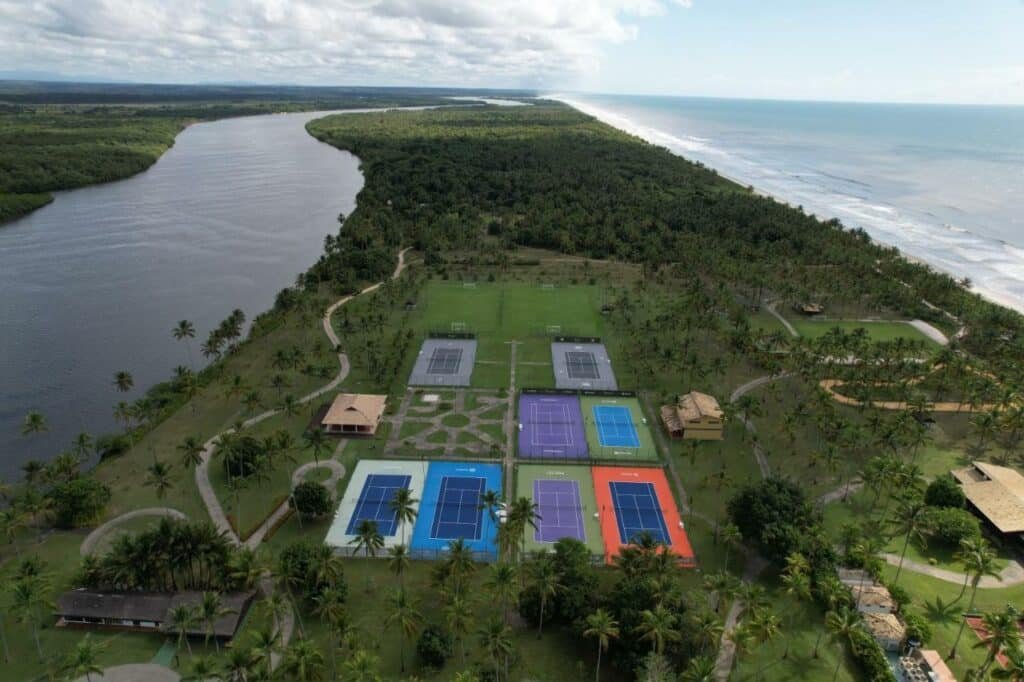 A imagem mostra uma vista aérea do Transamerica Comandatuba All Inclusive Resort, destacando a área de esportes do resort. Na parte inferior da imagem, várias quadras de tênis coloridas são visíveis, com cores como azul, roxo e laranja. Ao redor das quadras, há vastas áreas verdes com palmeiras, conferindo um ambiente tropical. À esquerda, o rio serpenteia ao longo da paisagem, enquanto à direita, a praia se estende paralelamente ao rio, criando um cenário natural contrastante entre a água doce e o mar. O céu está parcialmente nublado, adicionando um toque de suavidade ao panorama.