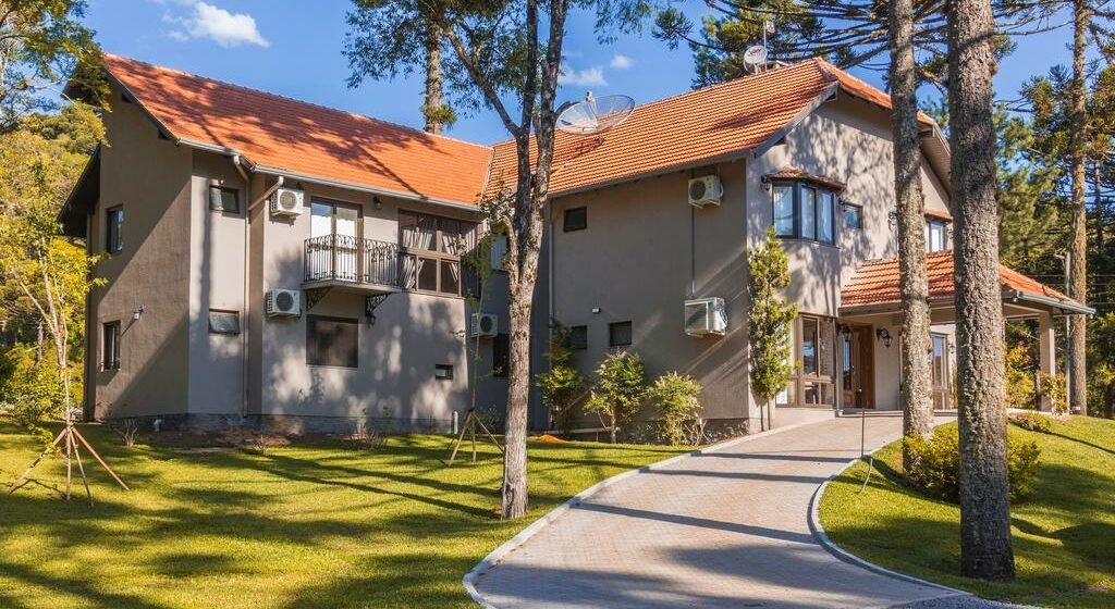 Uma casa cinza de dois andares na Tri Pousada Caracol com telhado de telhas vermelhas, cercada por árvores e um gramado bem cuidado, com um caminho que leva à entrada.