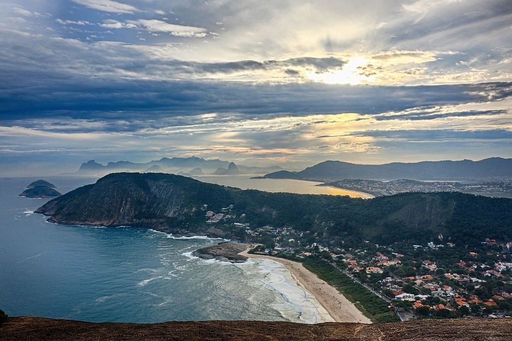 Vista aérea de uma paisagem costeira com praias, colinas e edifícios espalhados sob um céu nublado com luz do sol aparecendo.
