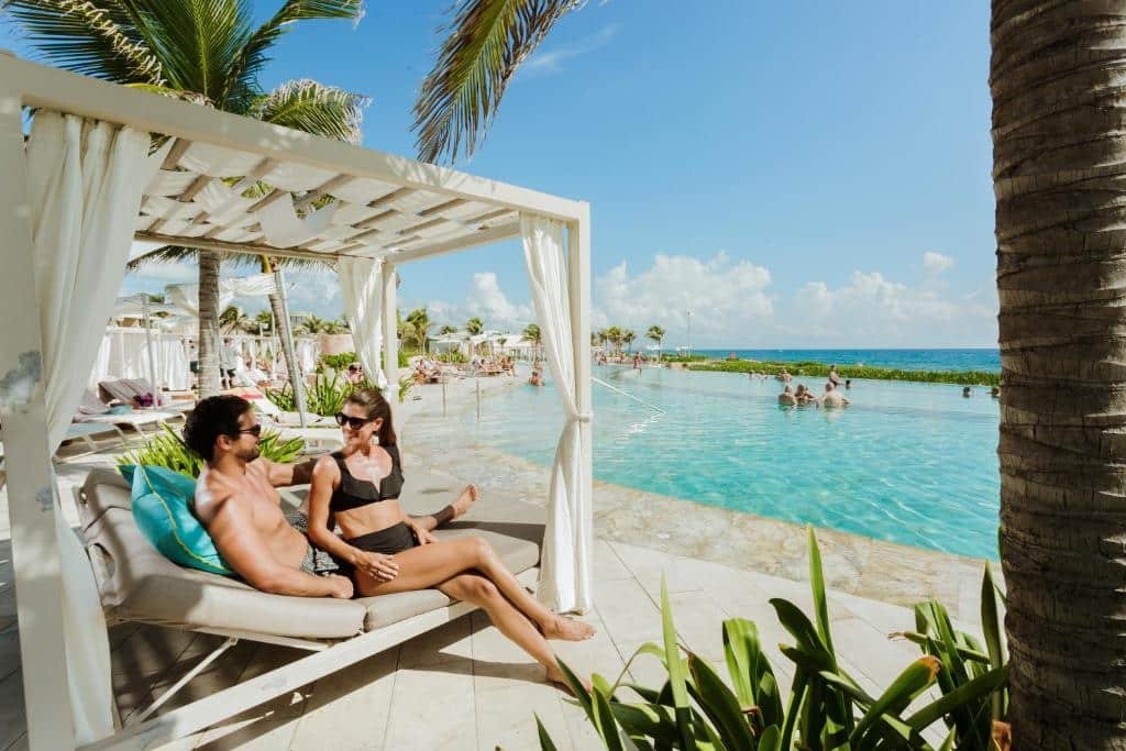 Um casal relaxa em uma espreguiçadeira sob um dossel perto de uma piscina. Palmeiras e pessoas na piscina são visíveis sob um céu azul claro para representar um dos melhores lugares para lua de mel