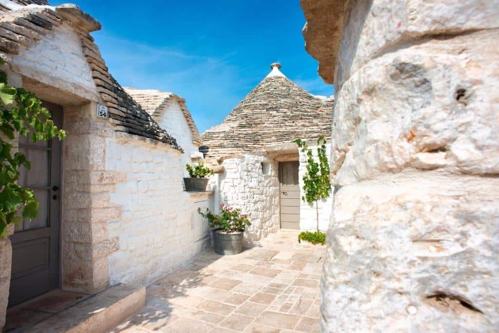 Fachada das entradas dos quartos do Trulli Holiday Albergo Diffuso com paredes de pedras brancas e telhados pontudos de pedras também. Na frente dos Trulli tem alguns vasos de plantas verdes. Imagem ilustrando post Hotéis em Puglia.