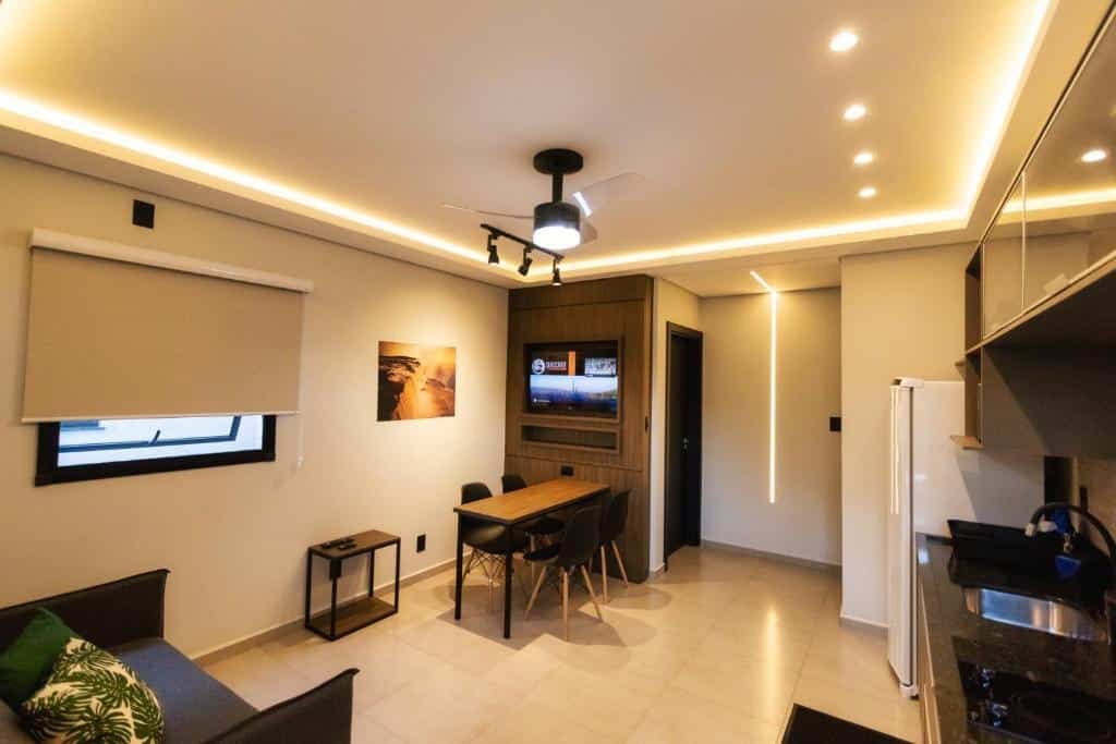 TuCasa Flats, e sua cozinha e sala de estar conjugada. A pia, armários, geladeira e outros eletrodoméstico estão à direita, e à esquerda, no canto inferior, há um sofá. Na frente do sofá há uma mesa com quatro cadeiras e um painel com televisão. O espaço é bem amplo.