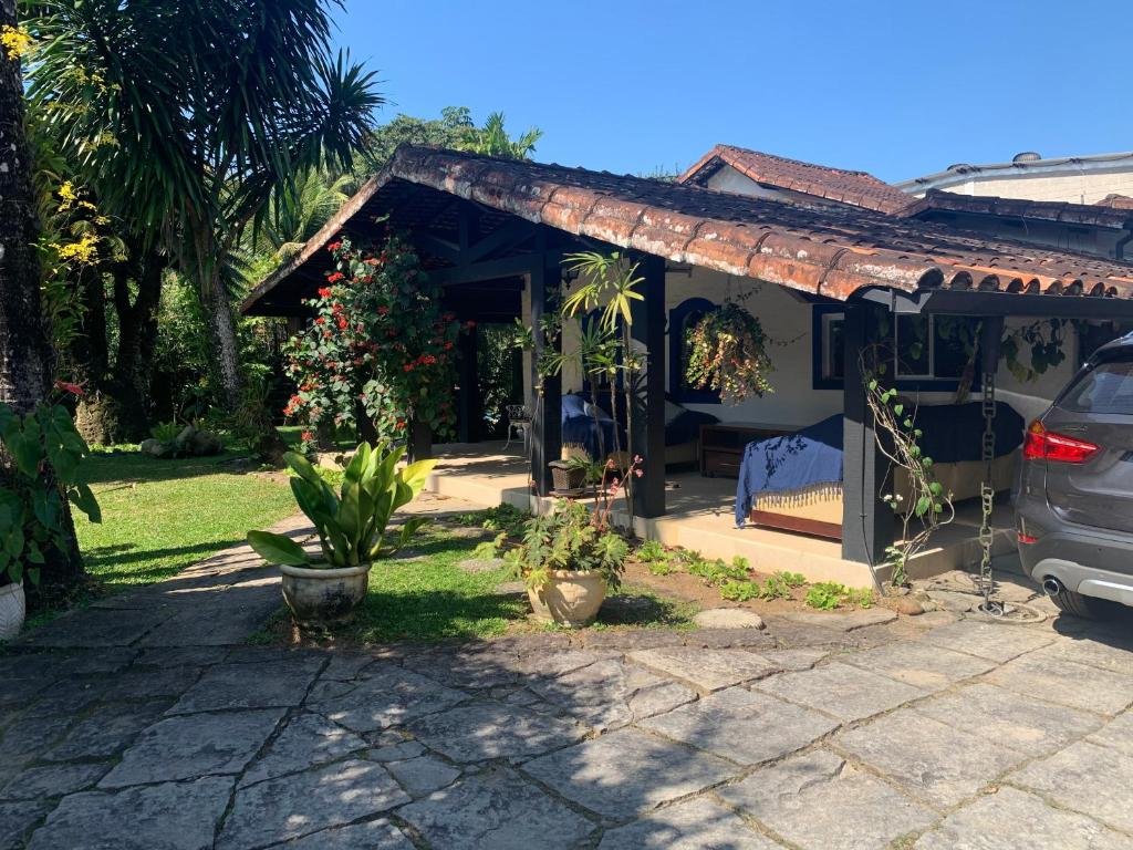 Área externa do Vivencie nossa energia. Do lado direito um carro, e a garage, da casa com sofás e vasos de planta. Do lado esquerdo uma caminho de pedra e o jardim da casa.
