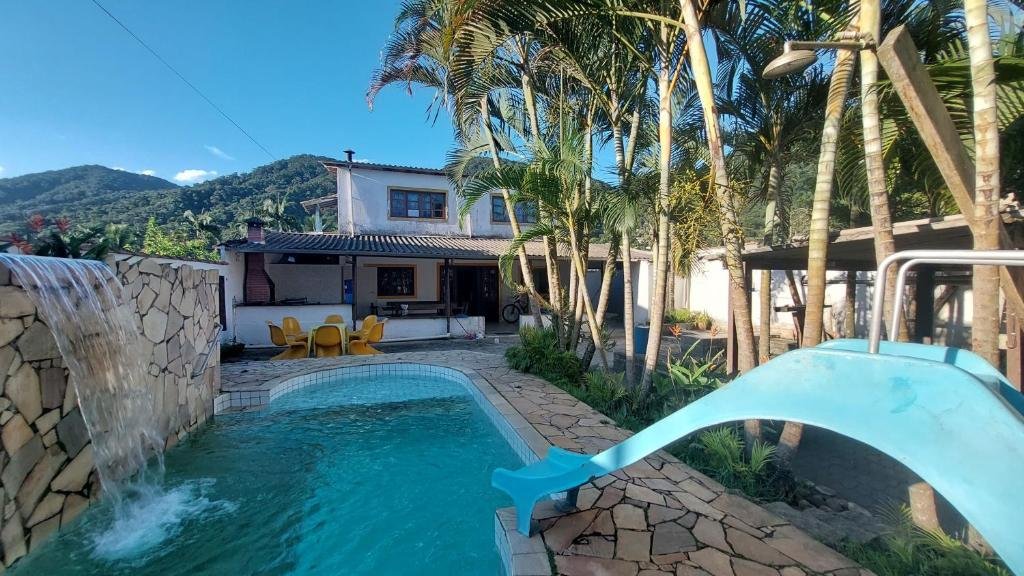 Área externa do Ubatubasul. Uma piscina na frente, com cascata e escorregador. No lado direito palmeiras e no fundo a casa.