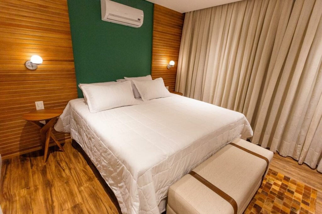 Cama de casal com colcha branca, recamier aos pés da cama, mesinha de cabeceira vazia, ar-condicionado, cortinas sobre a parede toda, e luzes de leitura