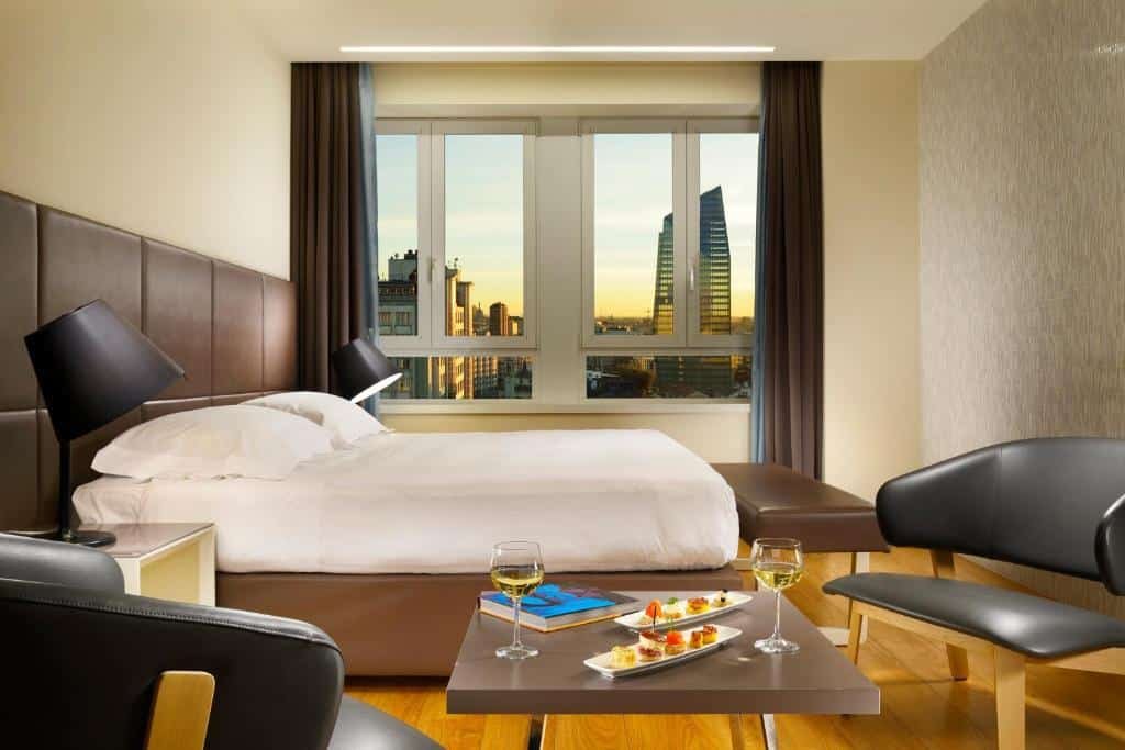 Quarto do UNAHOTELS Century Milano com uma janela com vista para a cidade, há uma cama de casal com uma cabeceira de courino marrom, ao pé da cama, há um pequeno banco do mesmo material, há uma pequena mesa quadrada com petiscos e vinho, e duas poltronas acolchoadas, nas laterais da cama, estão mesinhas de cabeceira com abajures em cima