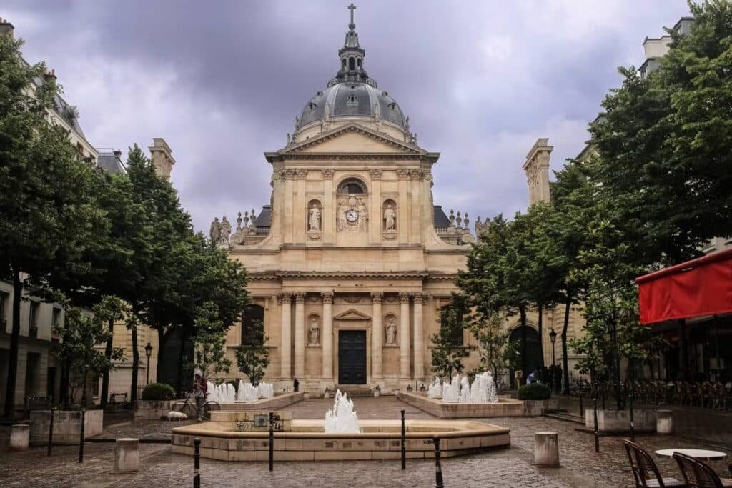 Um edifício histórico com telhado abobadado e torre do relógio, cercado por árvores, com uma fonte em primeiro plano sob um céu nublado. Essa é a Universidade de Sorbonne, uma das paradas num dos passeios em Paris.