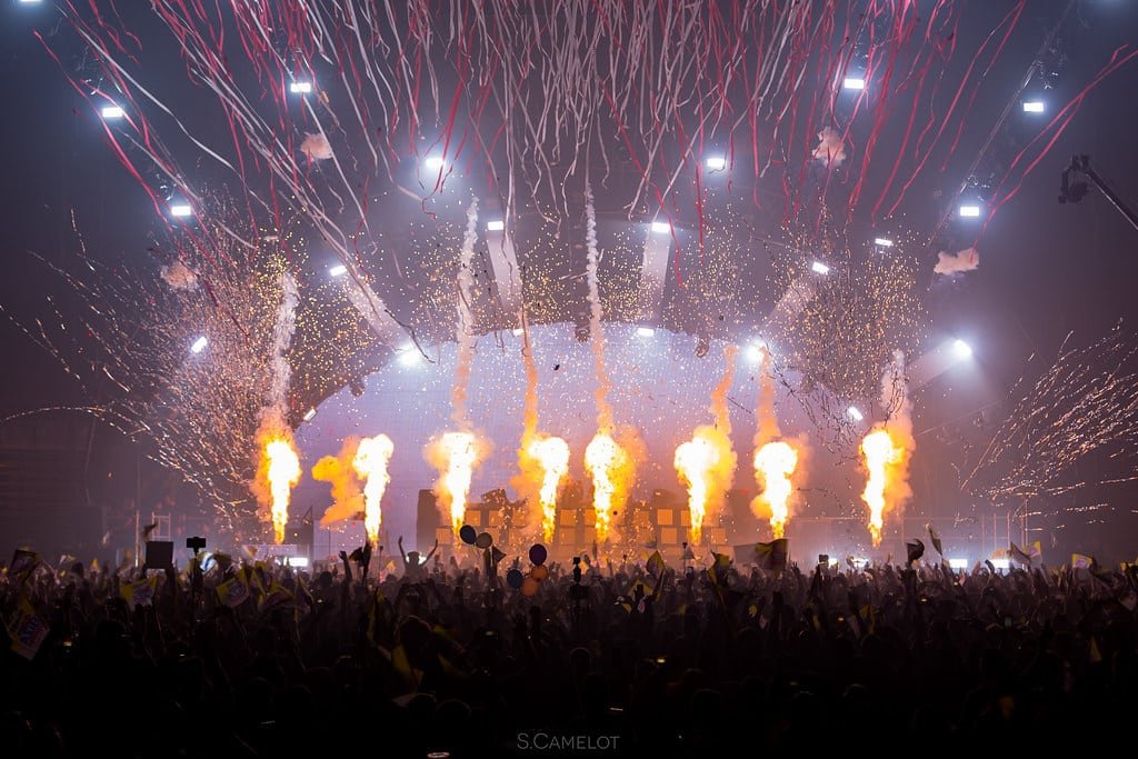 Uma grande multidão em um show com fogos de artifício e chamas no palco, cercada por serpentinas coloridas e luzes brilhantes no alto. Esse é o Ushuaïa, uma das festas em Ibiza.