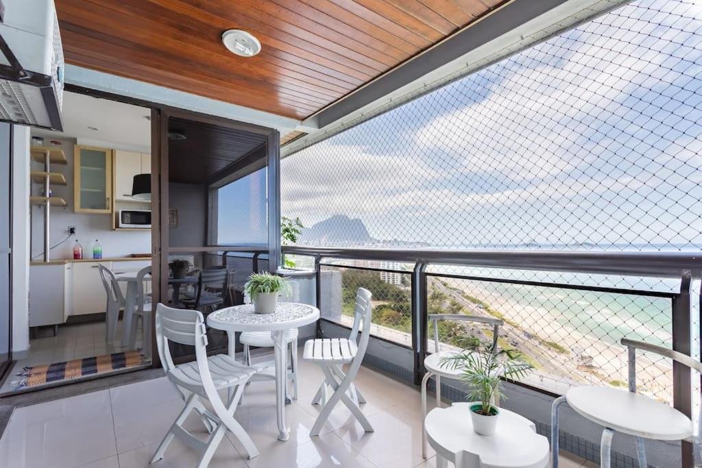 Varanda do Vista DE CINEMA do 19 andar da Praia BARRA da TIJUCA – Portaria 24h, Estacionamento, Wi-Fi 35mbps, Ar Condicionado – BANHEIRO RECEM REFORMADO durante o dia com duas cadeiras do lado direito da imagem logo a frente uma mesa redonda com três cadeiras.