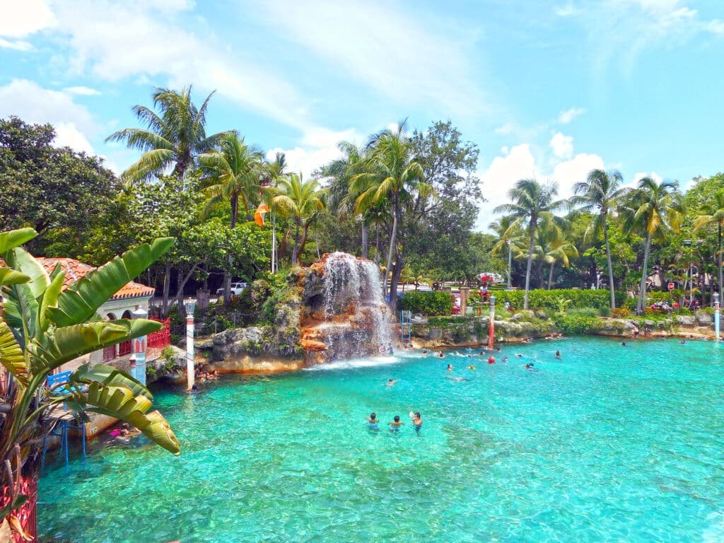Pessoas nadando em Venetian Pool. A piscina é bem grande e redonda e parece um rio com água bem azul. Algumas pedras e cachoeiras estão ao redor, assim como plantes e árvores.