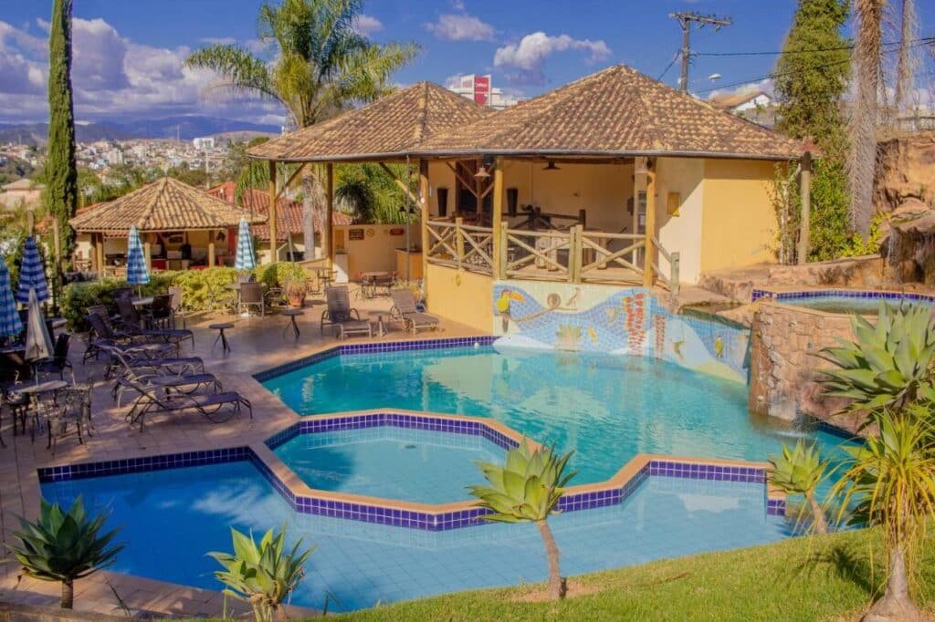 Área ao ar livre da Vila Chico com piscina de azulejos, espreguiçadeiras, guarda-sóis e uma estrutura sombreada com assentos de bar cercada por vegetação. Esse é um dos hotéis fazenda em Minas Gerais.