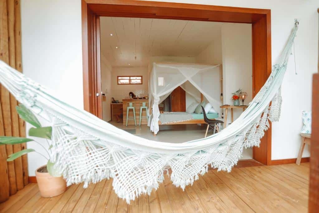 Vemos o interior e exterior do Villa Estrela Bungalow. Há uma varanda com rede de descanso no centro da foto, e no fundo vemos dentro do quarto. Há uma cama box, escrivaninha banheiro e cozinha equipada. Representa o post sobre airbnb em Itacaré.