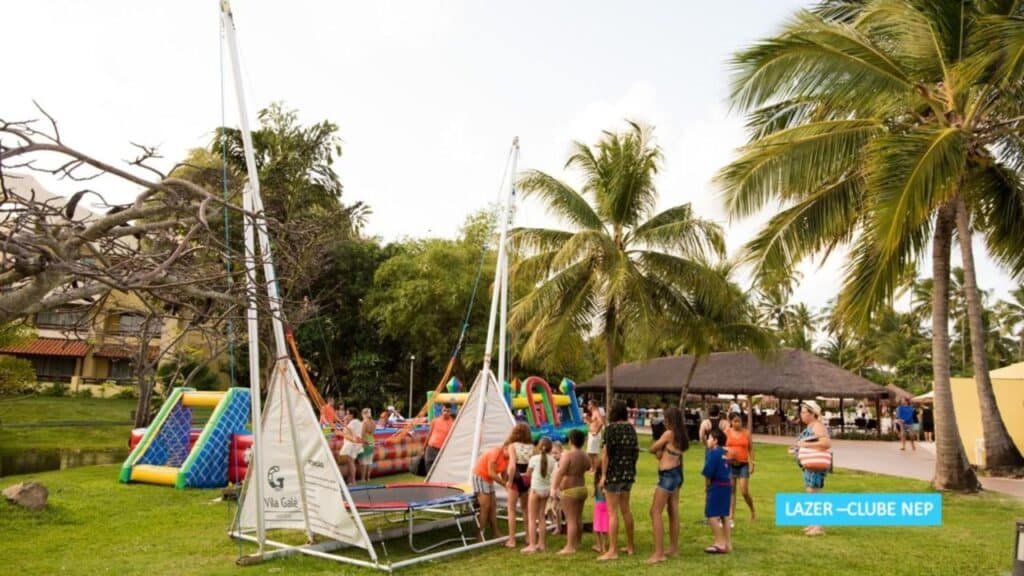 Pessoas se reúnem ao redor de um trampolim com arreios em um parque com estruturas infláveis e palmeiras ao fundo na Vila Galé Eco Resort do Cabo - All Inclusive.