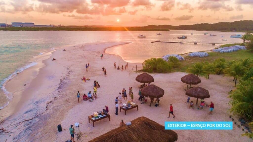 Uma praia ao pôr do sol com pessoas reunidas em torno de mesas e guarda-sóis de palha, com barcos na água e uma costa distante na Vila Galé Eco Resort do Cabo - All Inclusive.