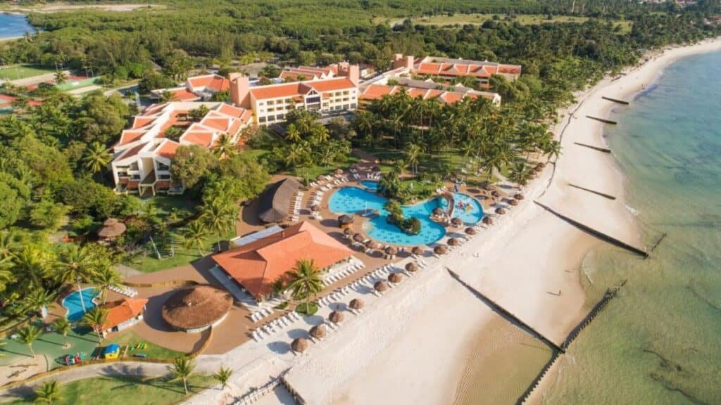 Vista aérea da Vila Galé Eco Resort do Cabo - All Inclusive à beira-mar com uma grande piscina, cercado por palmeiras e edifícios com telhados vermelhos.