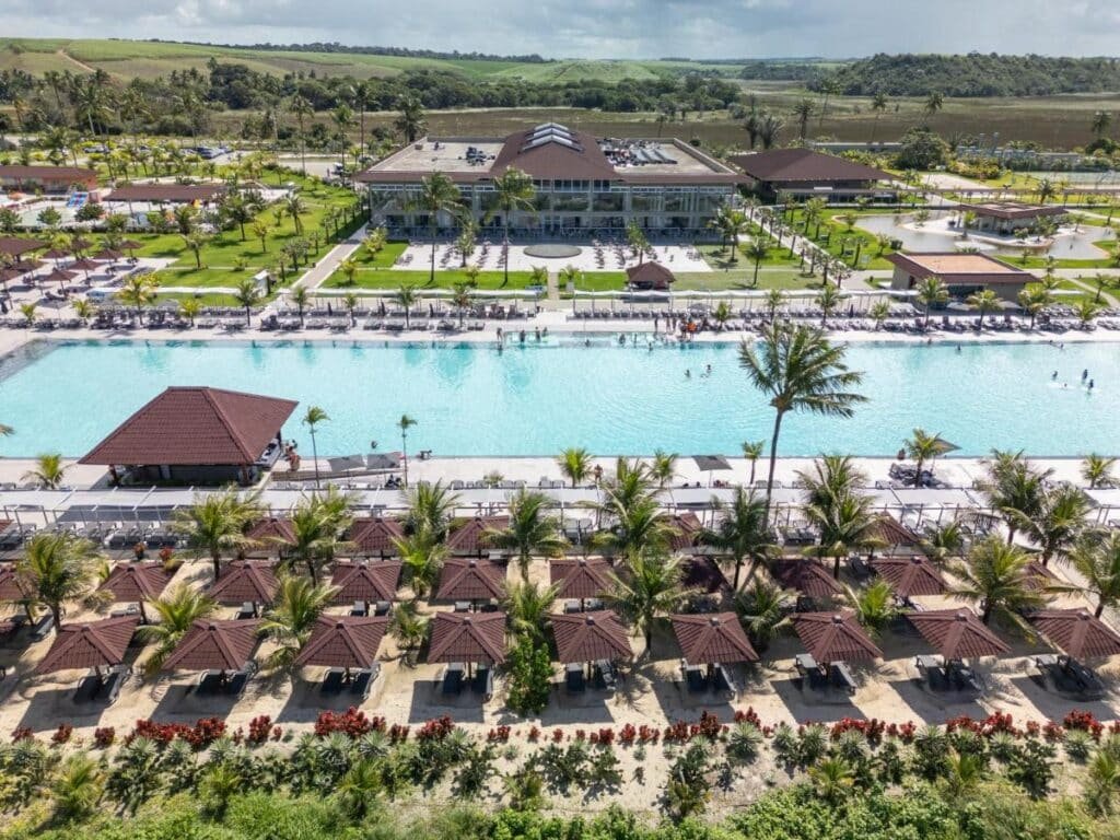 Vista aérea da Vila Galé Resort Alagoas - All Inclusive com uma grande piscina, cabanas e palmeiras em uma área paisagística, cercada por colinas verdes.