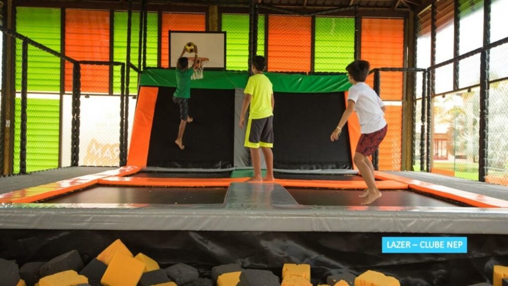 A imagem mostra três crianças brincando em uma área de trampolins dentro do Clube NEP no Vila Galé Resort Cumbuco, um dos melhores resorts all inclusive no Brasil. A estrutura é cercada por redes de proteção, com painéis coloridos nas paredes. Uma das crianças está pulando para enterrar uma bola de basquete na cesta, enquanto as outras duas observam e aguardam sua vez. No chão, há blocos de espuma em amarelo e preto para amortecer quedas, garantindo a segurança durante as brincadeiras.