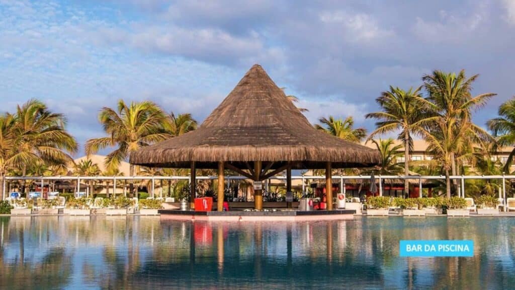 A imagem mostra um bar de piscina no Vila Galé Resort Cumbuco, um dos melhores resorts all inclusive no Brasil. O bar tem um telhado de palha em formato cônico e está localizado no centro da piscina, com acesso direto pela água. Ao fundo, palmeiras e estruturas do resort complementam a paisagem, que é iluminada pela luz suave do início da manhã ou final da tarde. O local transmite uma atmosfera relaxante e tropical, ideal para aproveitar bebidas refrescantes à beira da piscina.