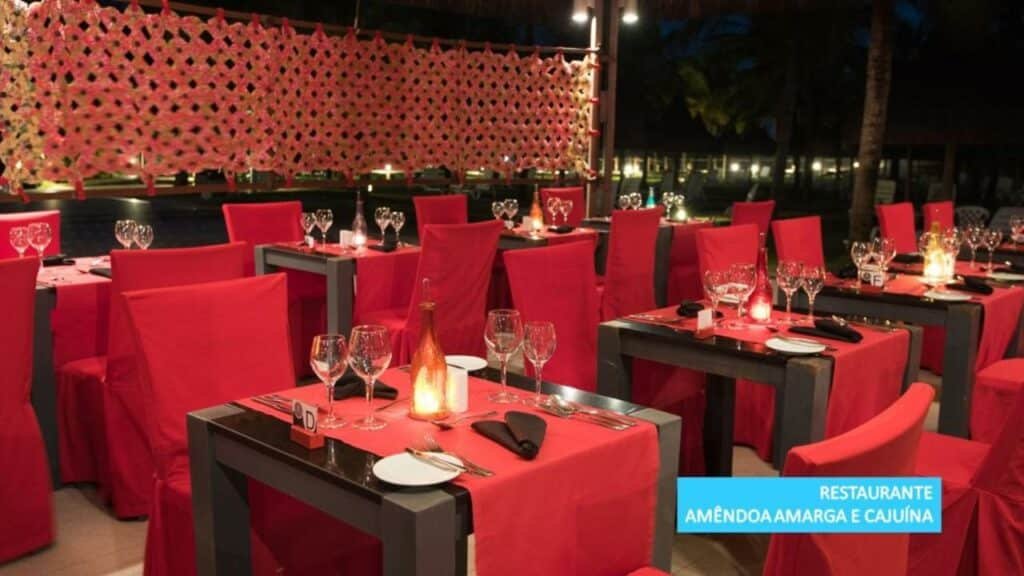 A imagem mostra o restaurante Amêndoa Amarga e Cajuína no Vila Galé Resort Cumbuco. O ambiente é elegante e acolhedor, com mesas quadradas cobertas por toalhas vermelhas e cadeiras combinando com capas vermelhas. As mesas estão arrumadas com talheres, pratos, copos de vinho e guardanapos pretos, prontos para receber os convidados. Há garrafas decorativas e velas acesas, que proporcionam uma iluminação suave e romântica ao local. Ao fundo, é possível ver uma decoração de tecido trançado e luzes que iluminam o ambiente, criando uma atmosfera charmosa e convidativa para o jantar.