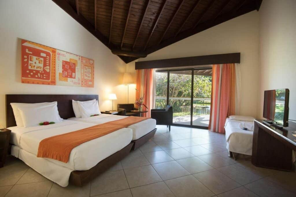 Quarto da Vila Galé Resort Marés - All Inclusive com duas camas, cor laranja, TV e uma grande janela com cortinas com vista para um jardim.