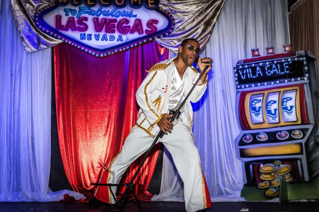 Artista vestido com um terno branco com detalhes dourados canta no palco perto de uma placa temática de Las Vegas e de um cenário de máquina caça-níqueis na Vila Galé Resort Marés - All Inclusive.