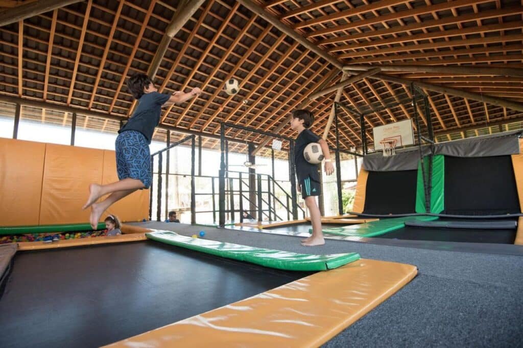 Duas crianças brincando com bolas de futebol em trampolins internos, sob um teto de madeira. Uma cesta de basquete é visível ao fundo.