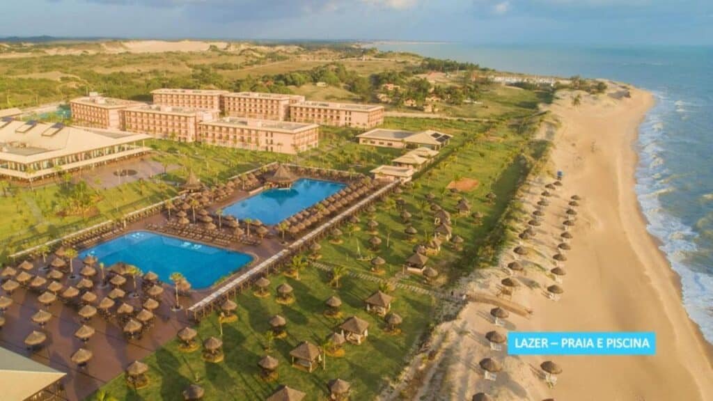 Vista aérea da Vila Galé Resort Touros à beira-mar com vários edifícios, uma grande piscina e guarda-sóis de palha em uma praia de areia.