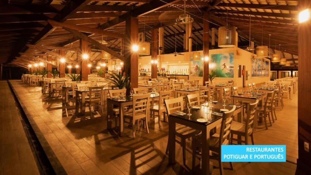 Um restaurante espaçoso na Vila Galé Resort Touros com mesas e cadeiras de madeira sob iluminação aconchegante, com uma grande área de bar ao fundo.