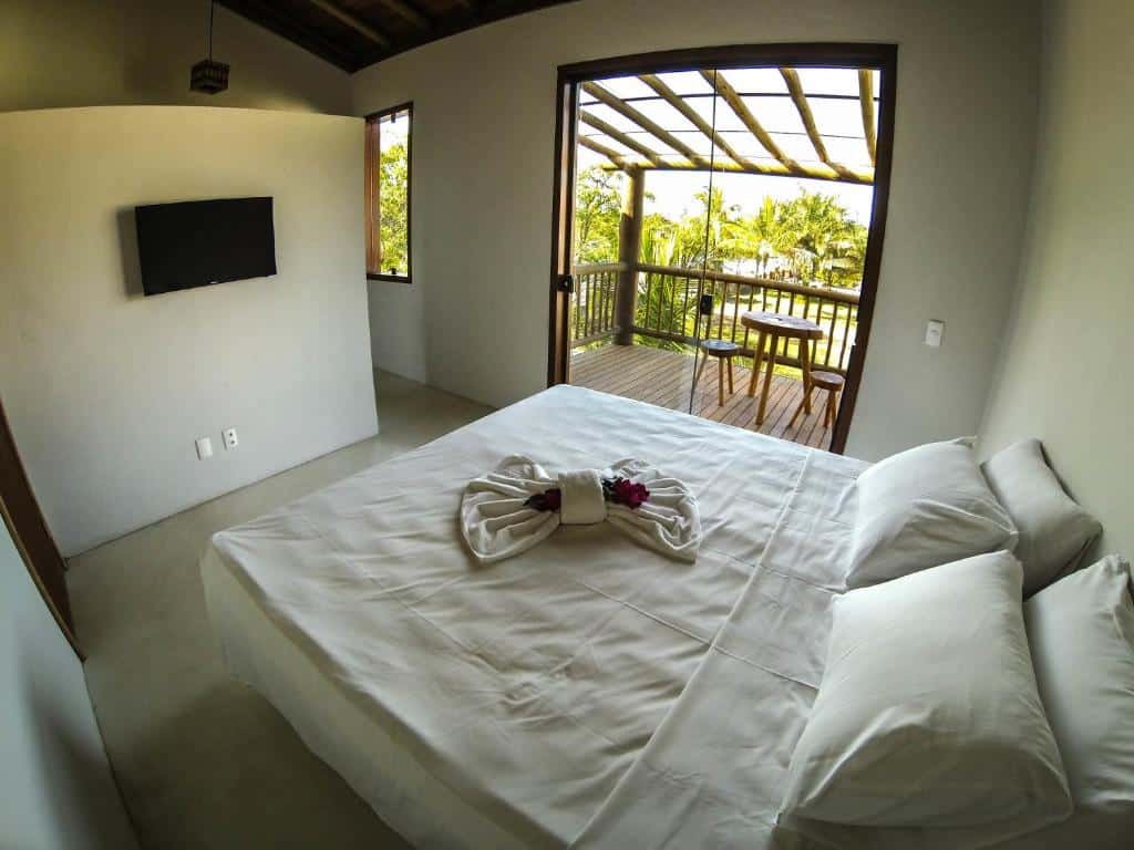 Vista de cima do quarto na Vila Pérola Negra. A cama é box e de casal e está à direita. Na frente há uma parede com televisão. Do lado esquerdo a cama há uma varanda com mesa e bancos. Ainda na direita, porém mais ao fundo, há uma porta. Representa o post sobre airbnb em Itacaré.