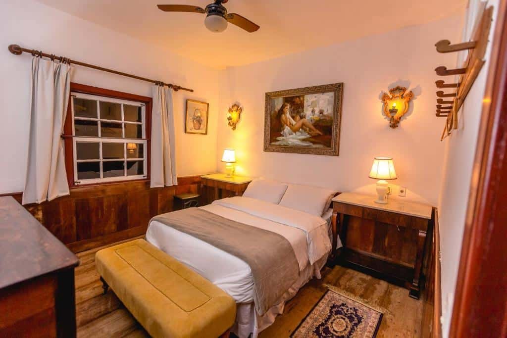 A imagem mostra um quarto aconchegante e elegantemente decorado do Hotel Vila Relicário. No centro, há uma cama de casal com lençóis brancos e uma manta cinza, em frente à cama há um banco estofado amarelo. As paredes brancas contrastam com o rodapé de madeira escura. Ao lado da cama, há dois criados-mudos com luminárias de abajur. Acima da cama, um quadro clássico emoldurado com a imagem de uma mulher decora a parede. O ambiente é iluminado por uma luz suave, e há uma janela com cortinas claras ao lado da cama, além de um ventilador de teto.